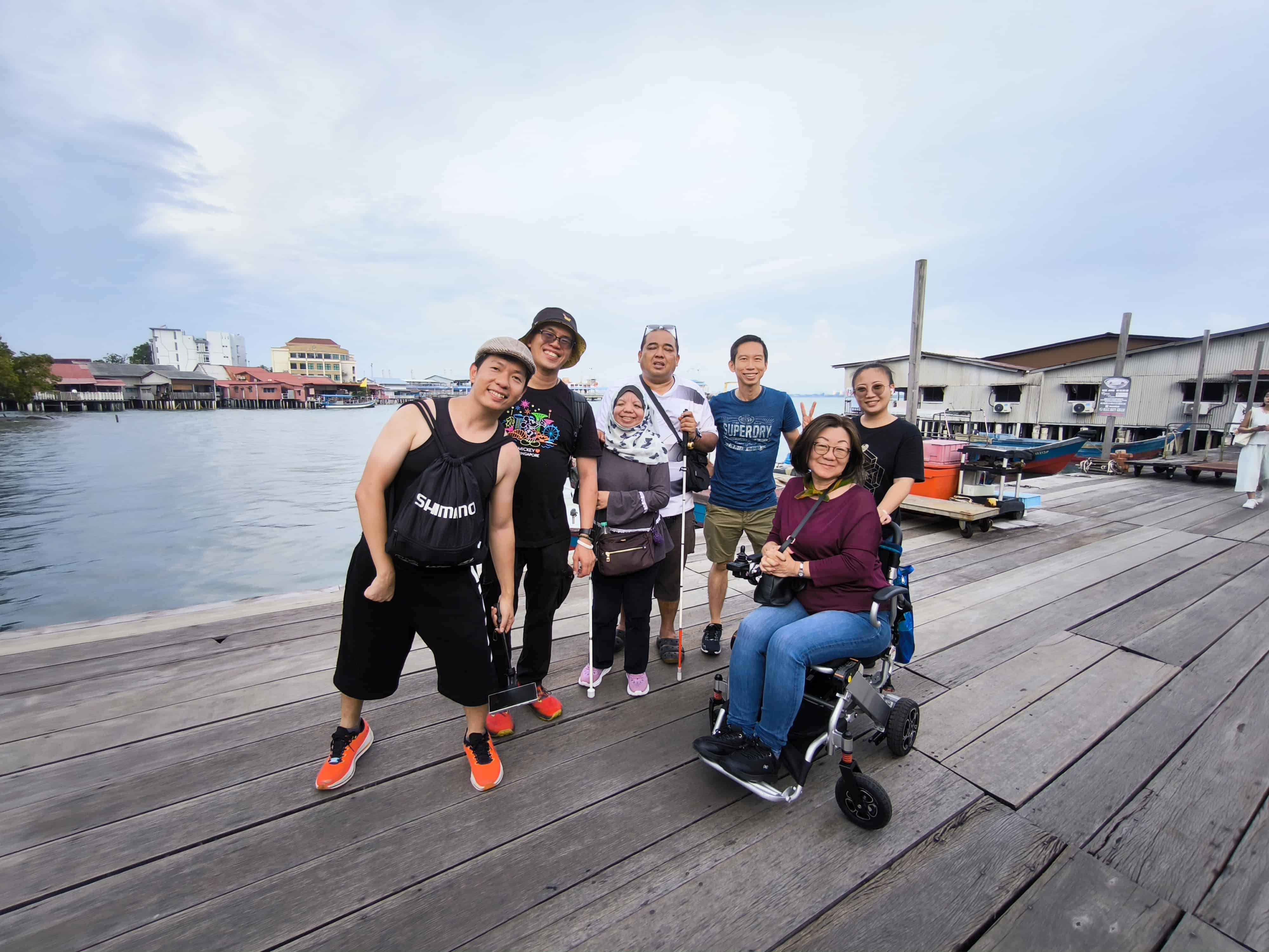 Group of travellers posing on the wooden boardwalk at Chew Jetty in Penang, with waterfront houses and boats in the background.