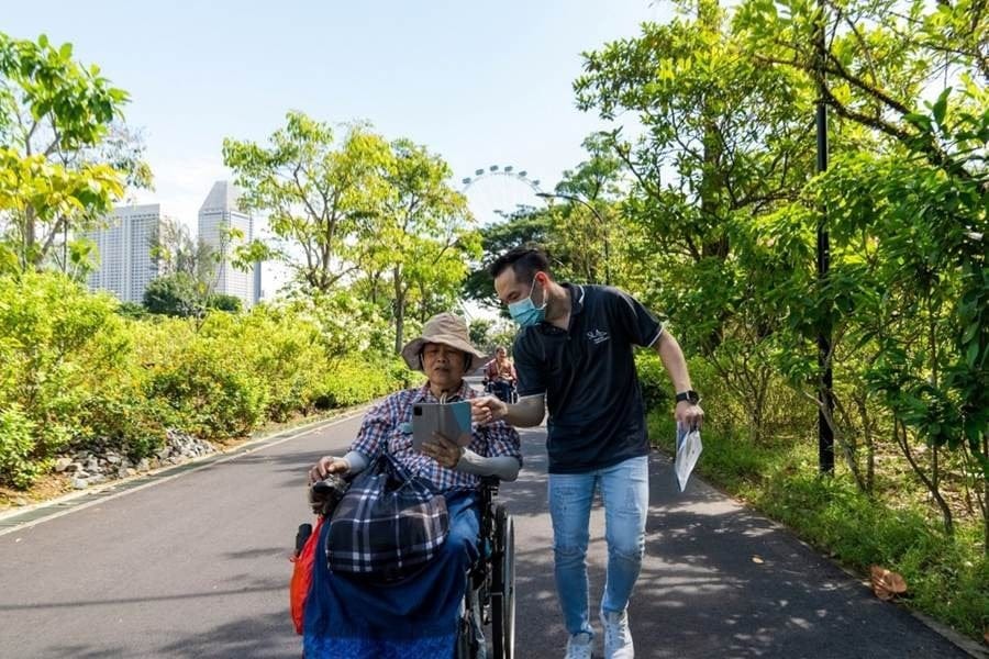 A guide shows information on a tablet to an older wheelchair user along an accessible garden path in Singapore. 