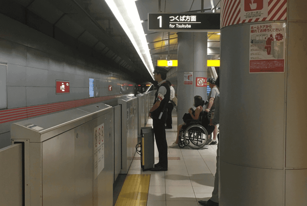 A wheelchair user waiting with other passengers beside a station attendant on an accessible train platform in Japan.