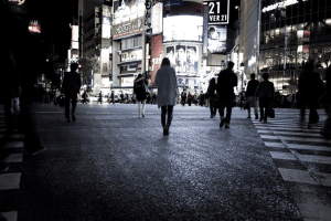 Pedestrians crossing a busy, brightly lit city intersection in Japan at night.