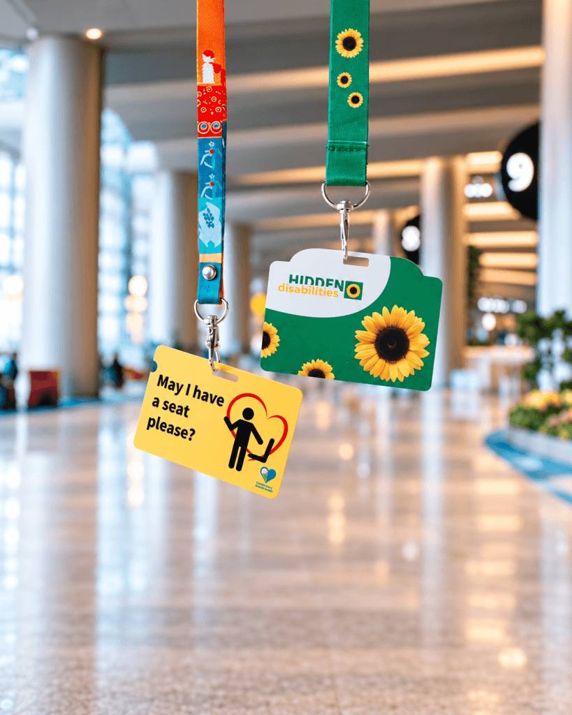 Two hidden-disability awareness lanyards displaying “Hidden Disabilities” and “May I have a seat please?” cards in a bright indoor public space.
