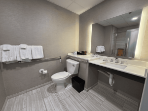 Accessible hotel bathroom with grab bars beside the toilet, wide floor space and an open area beneath the sink for wheelchair access.