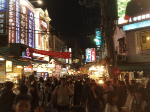 Crowds walking among brightly lit shops and food stalls at a busy night market in Taiwan.