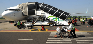 Airport staff assisting a wheelchair user across the tarmac toward an aircraft fitted with a covered