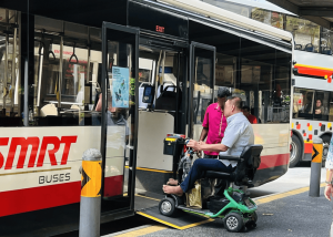 A mobility scooter user boarding an accessible public bus using a wheelchair ramp, assisted by a staff member.
