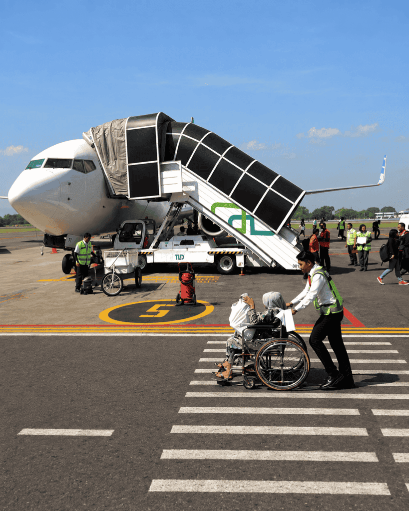 An airport staff member assists a wheelchair user across the tarmac in front of a passenger aircraft and covered boarding stairs.