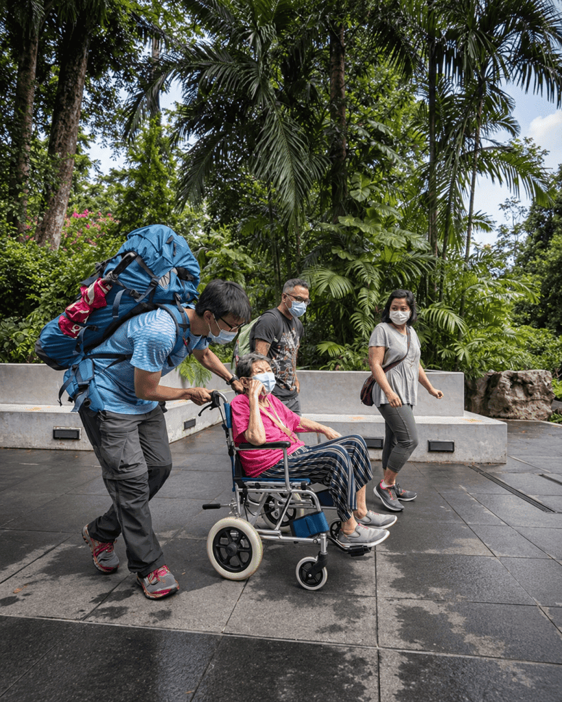 A man pushes an older woman in a wheelchair along an accessible path through a tropical garden, accompanied by two other visitors.