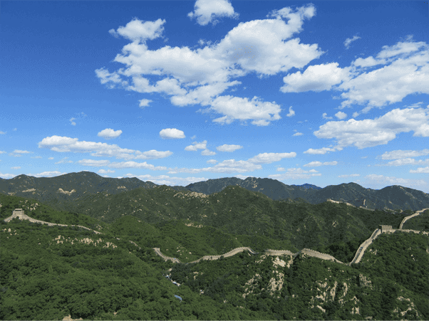 Great Wall of China winding across green mountain ridges beneath a blue sky with scattered clouds.