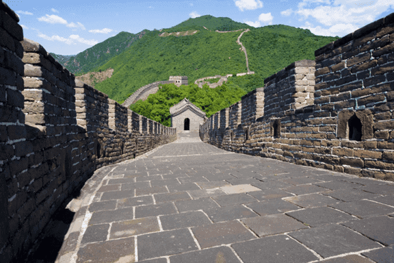 Stone walkway along the Great Wall of China, bordered by brick walls and leading towards a watchtower and green mountains.