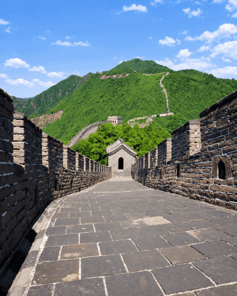 Stone walkway along the Great Wall of China, bordered by brick walls and leading towards a watchtower and green mountains.